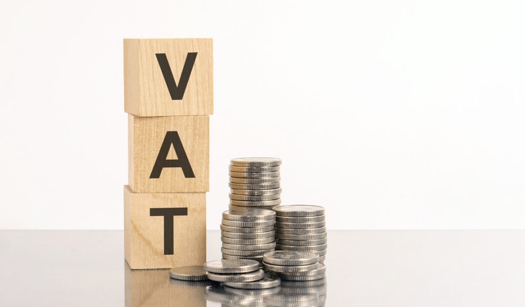 the word VAT is laid out of wooden cubes with letters on white background with coins. VAT - short for Value Added Tax